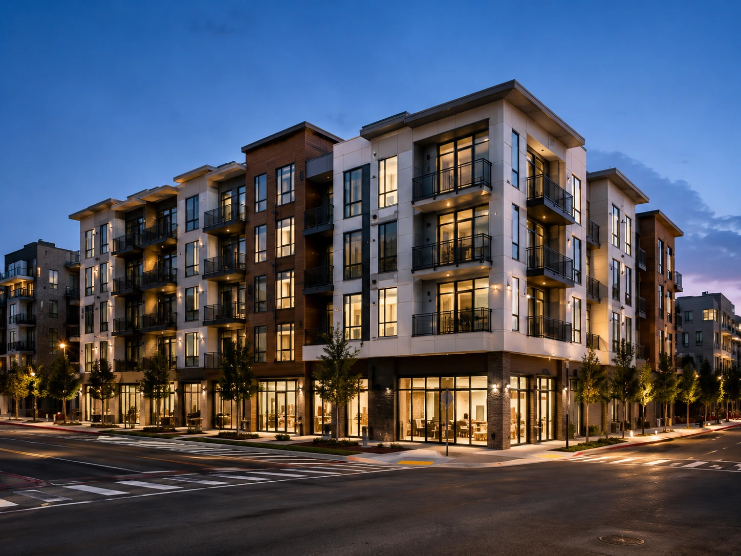Four-story modern apartment building with large windows and balconies, located at a street corner, illuminated at dusk.